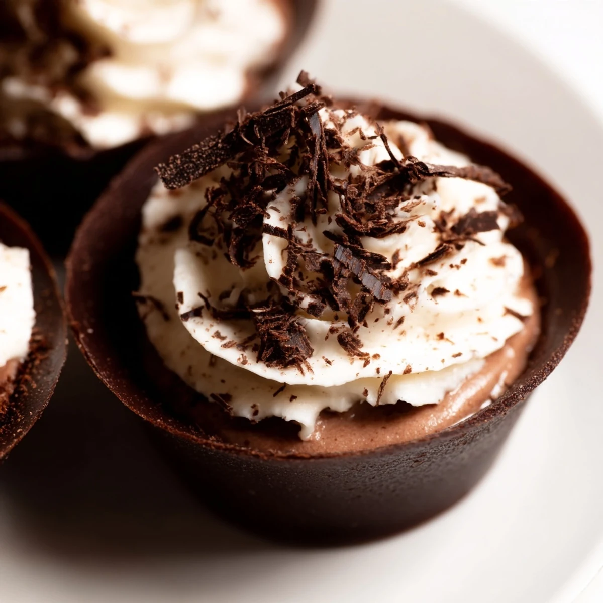 Four glossy, dark chocolate cups filled with rich chocolate mousse, crowned with soft whipped cream and chocolate shavings on a slate serving plate.