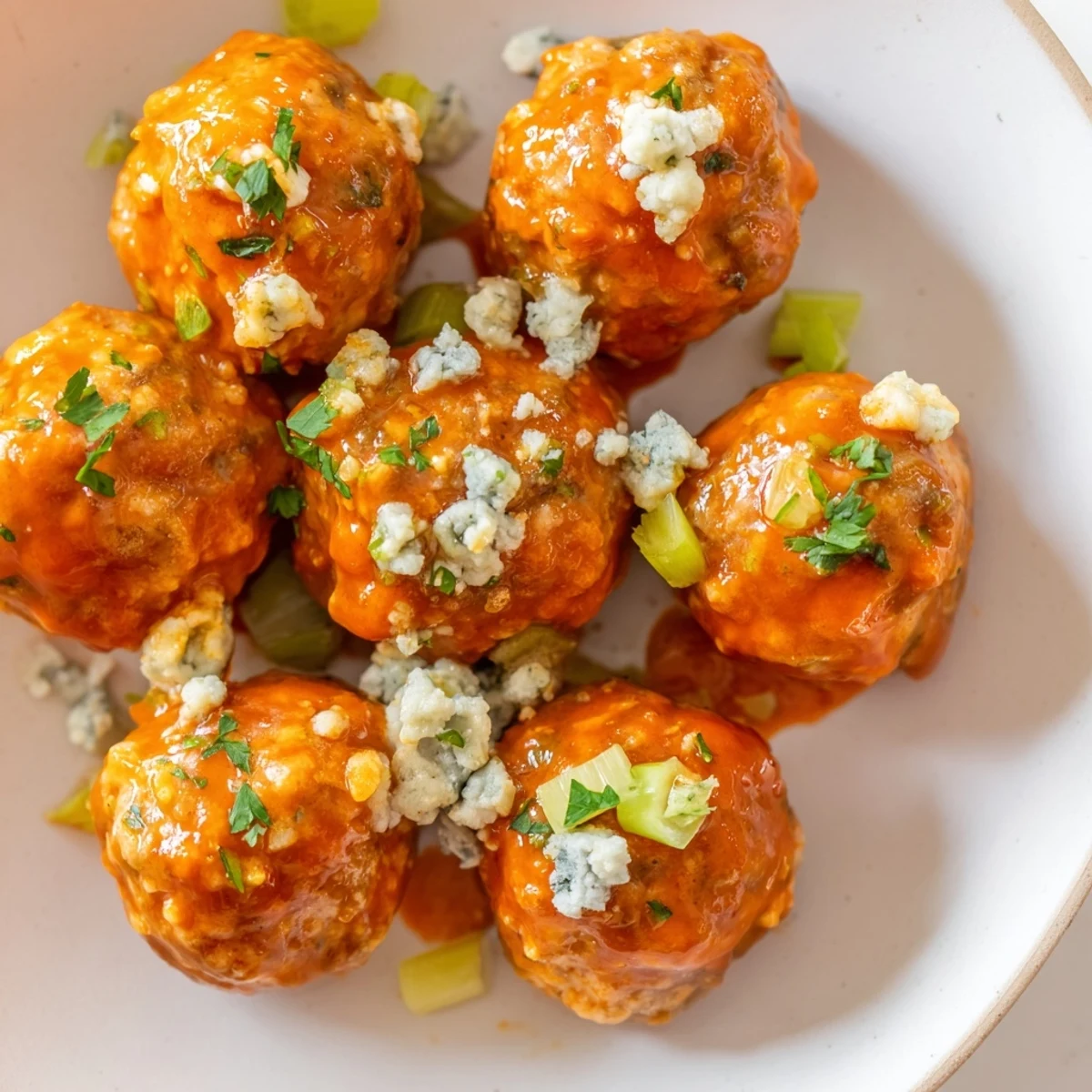 A close-up view of juicy Game Day Spicy Buffalo Chicken Meatballs, garnished with blue cheese crumbles and served with crisp celery sticks.