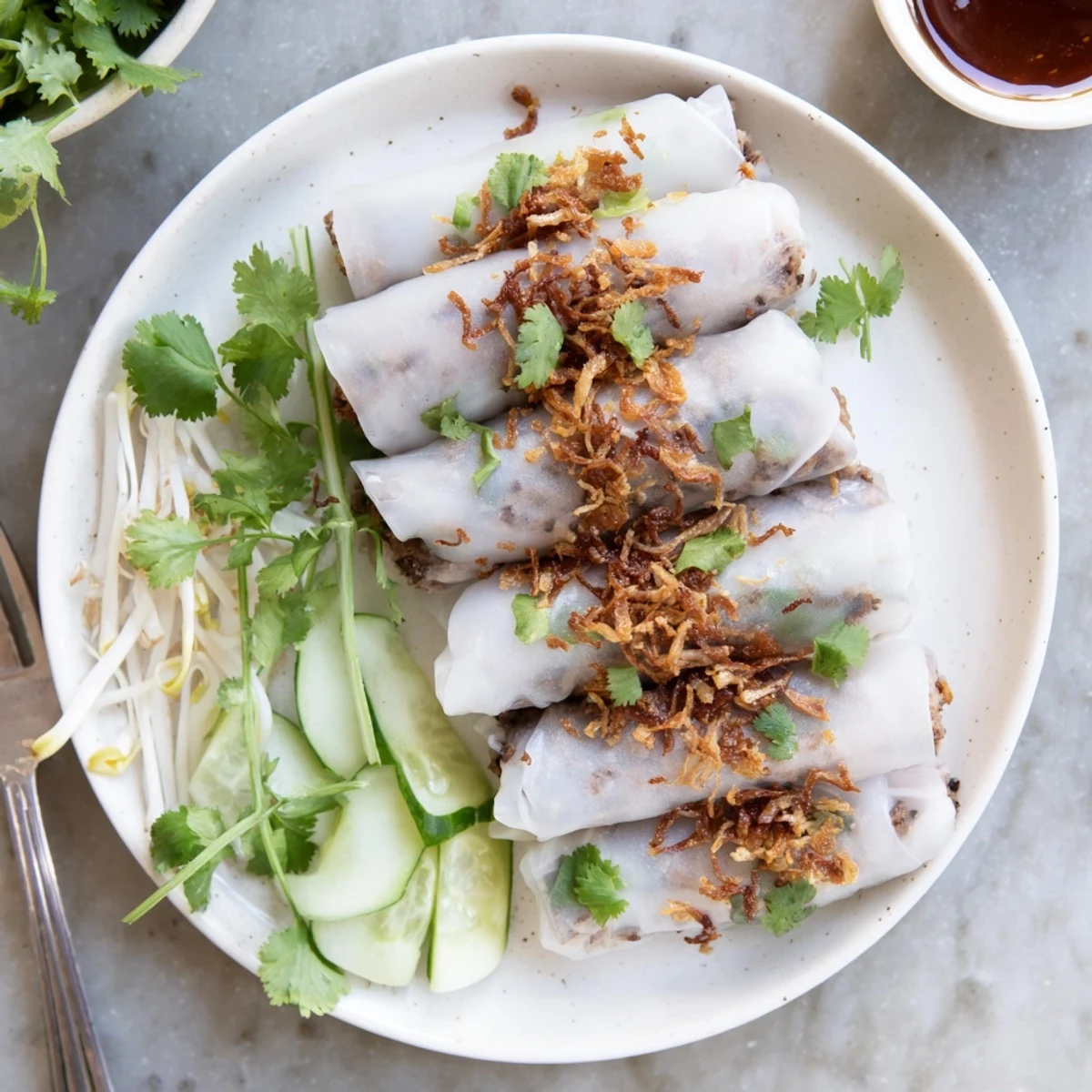 Steamed Banh Cuon rice rolls filled with savory pork and wood ear mushrooms, topped with fresh herbs, fried shallots, and cucumber slices.