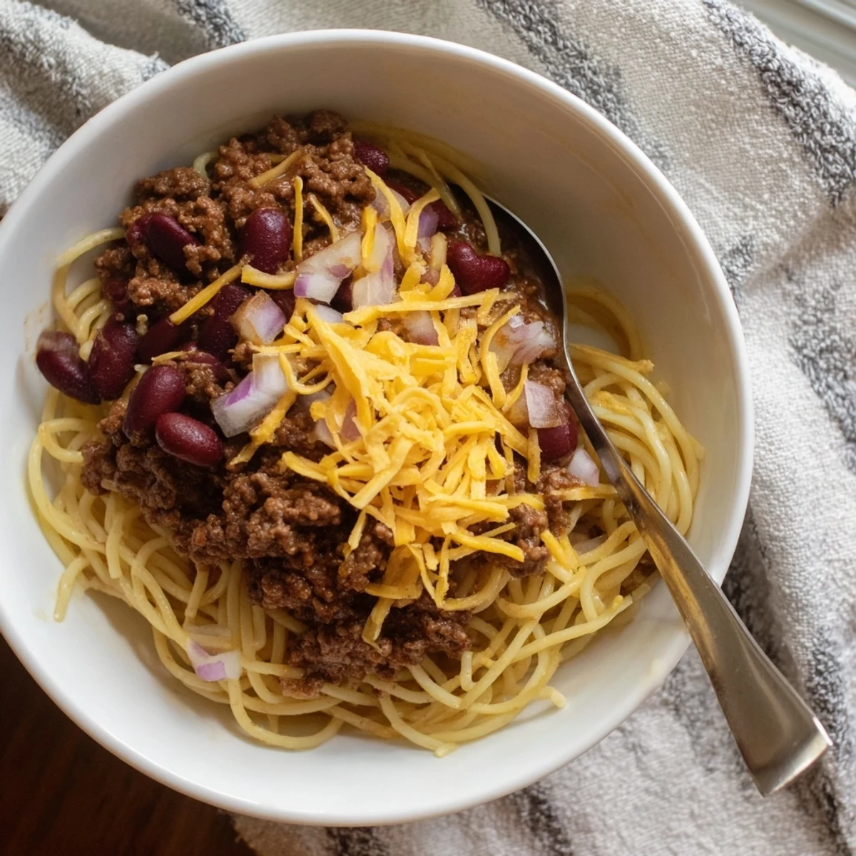 Golden Cincinnati Chili ladled over spaghetti with melted cheddar cheese and diced onions