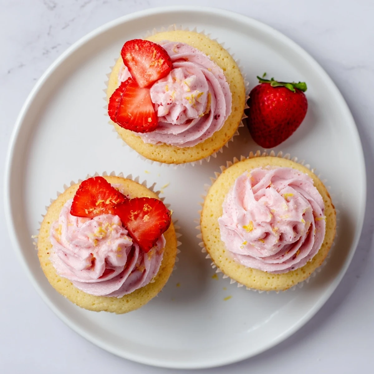 Strawberry lemonade cupcakes topped with pink strawberry frosting and fresh strawberry halves on a white plate