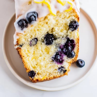 Overhead view of a Lemon Blueberry Yogurt Loaf Cake on a rustic wooden board, surrounded by fresh blueberries and lemon slices for a vibrant, summery presentation.  